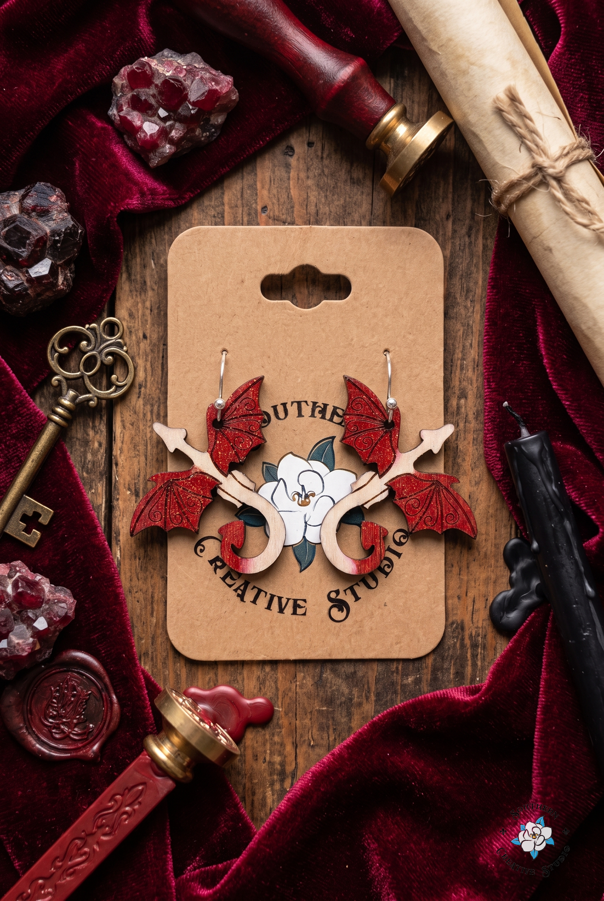 Flat lay of red dragon earrings on SCS card with crimson velvet, garnet, wax seals, key and parchment scroll