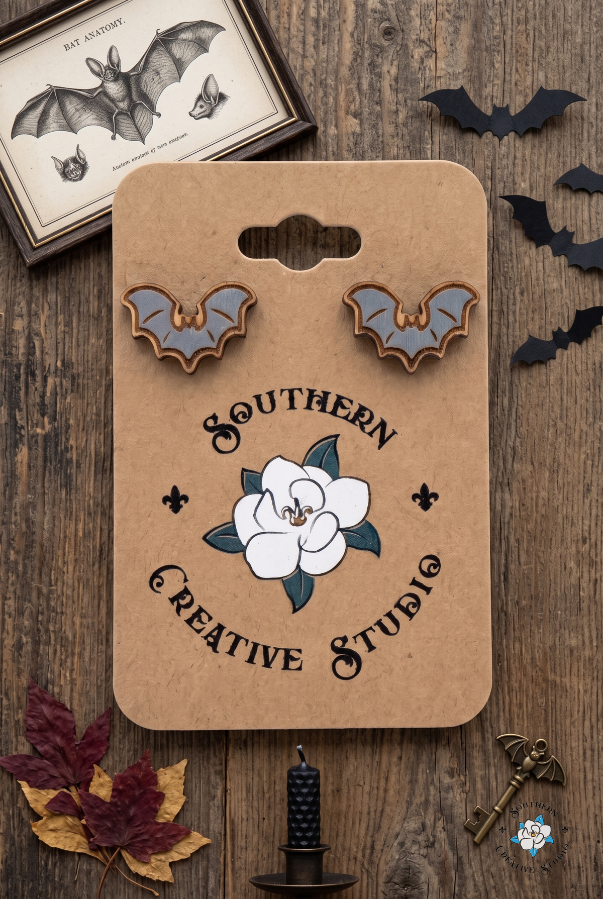 Flat lay of gray bat stud earrings on SCS card with bat print, paper bats and candle