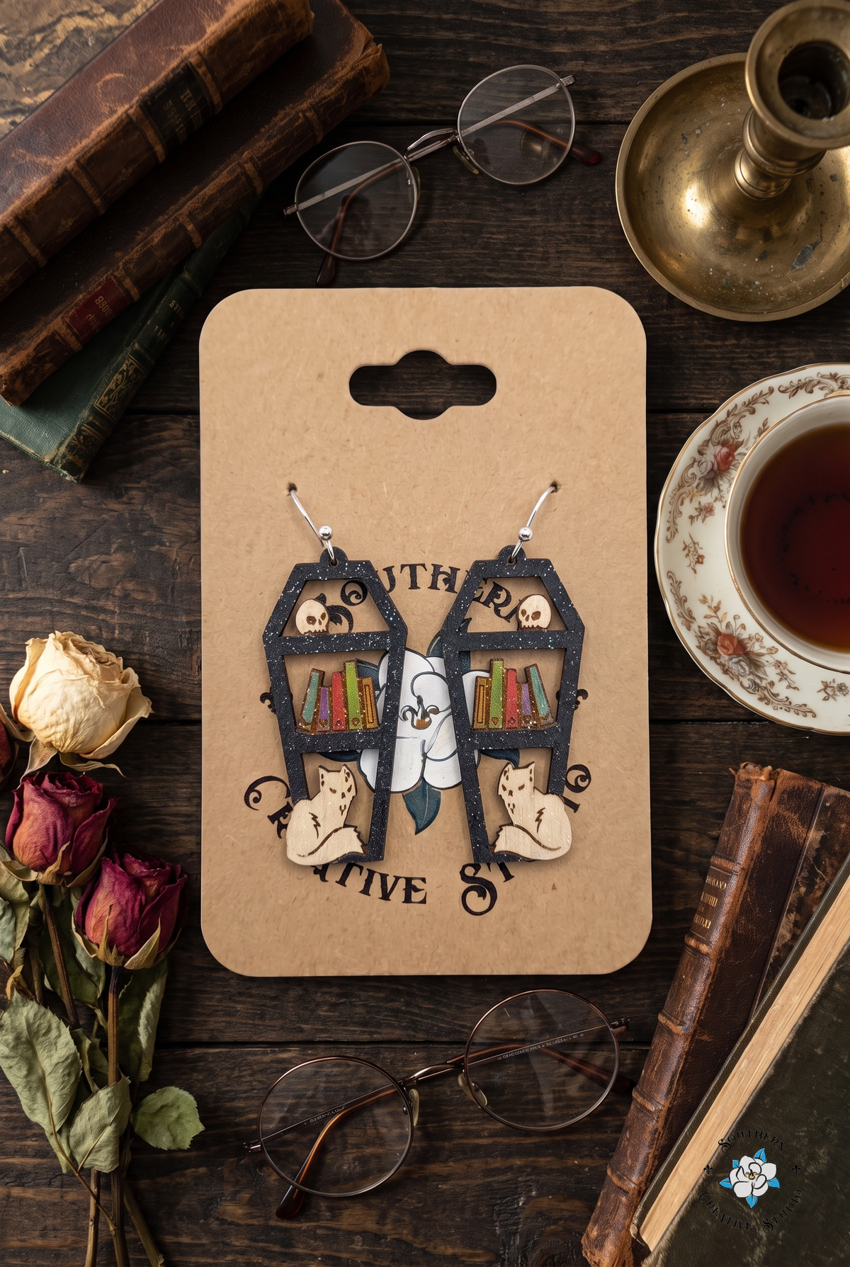 Flat lay of coffin bookshelf earrings on SCS card with old books, glasses and tea