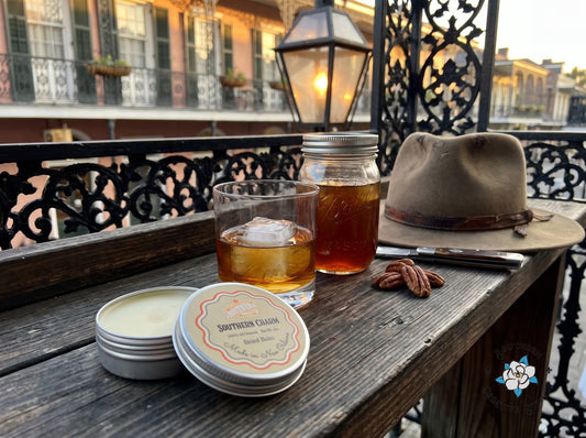 Southern Charm beard balm by Southern Creative Studio, open tin and lid with bourbon, a felt hat and a pocketknife on a New Orleans balcony rail