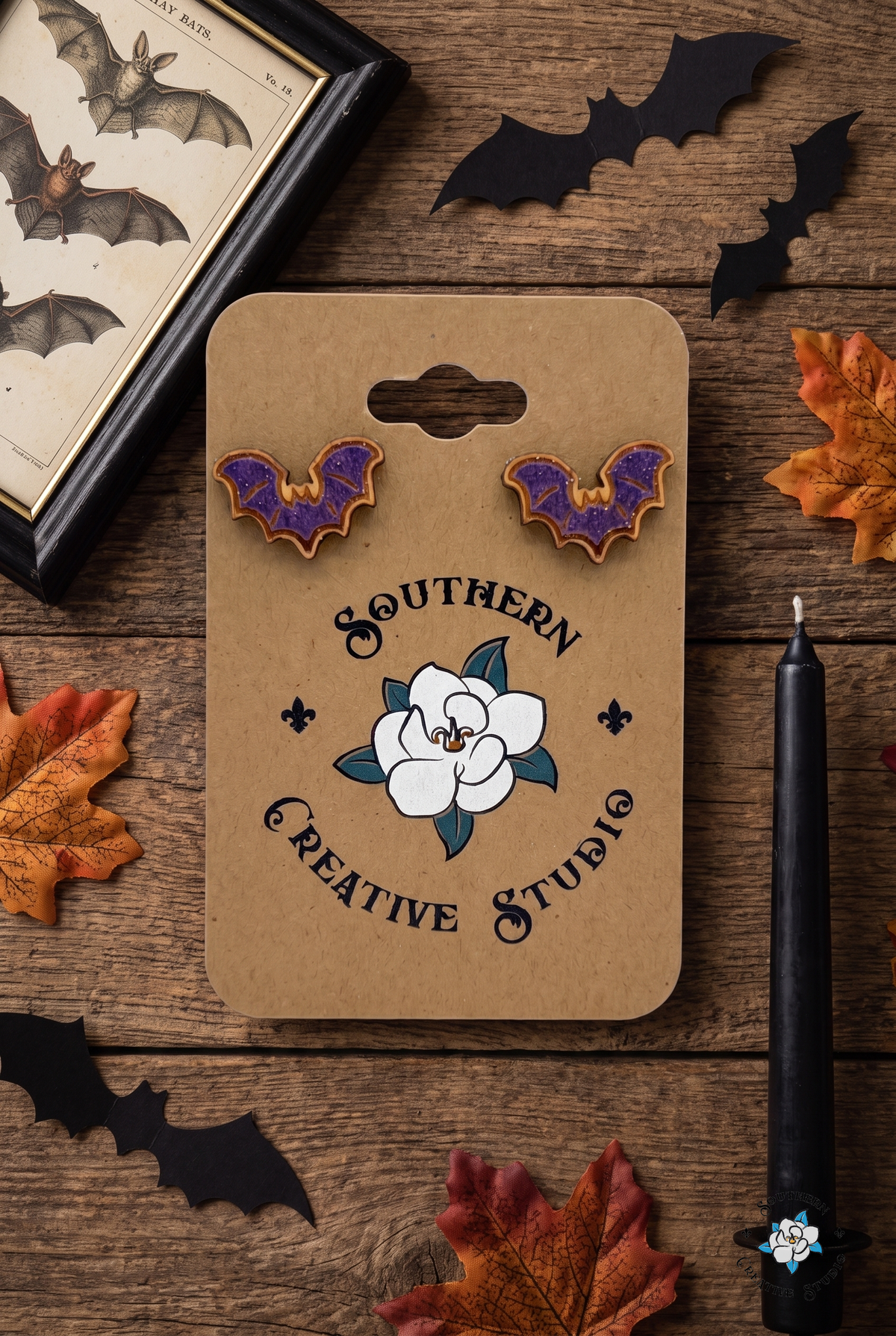 Flat lay of purple bat stud earrings on SCS card with bat print, paper bats and candle