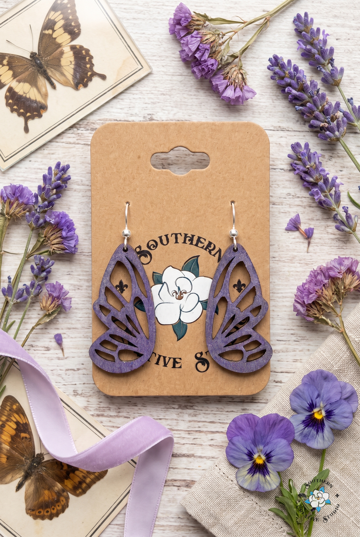 Flat lay of purple butterfly wing earrings on SCS card with lavender, pansies, lilac ribbon and butterfly specimen cards