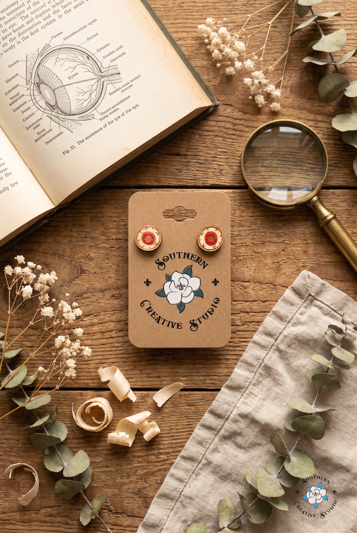 Red eyeball stud earrings on card flat lay with anatomy book and eucalyptus