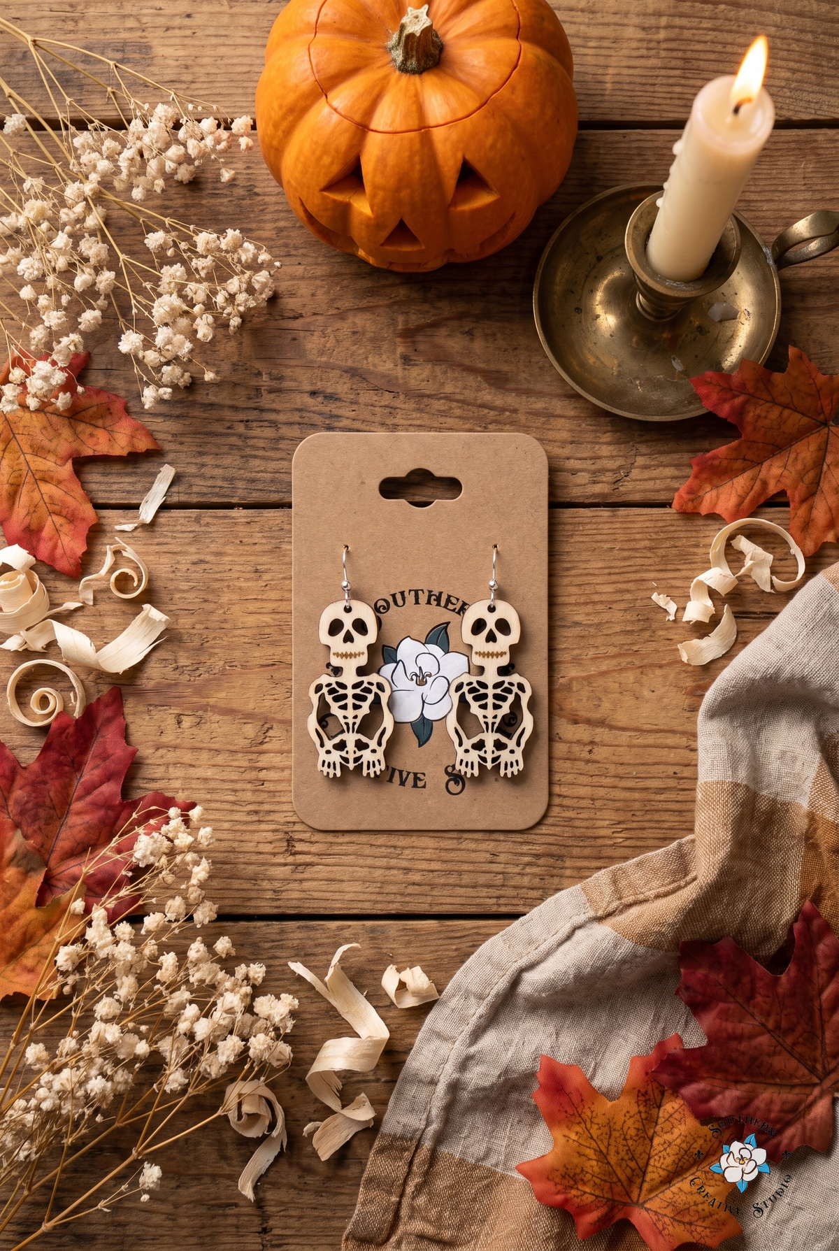 Skeleton earrings on card flat lay with Halloween pumpkin, candle and autumn leaves