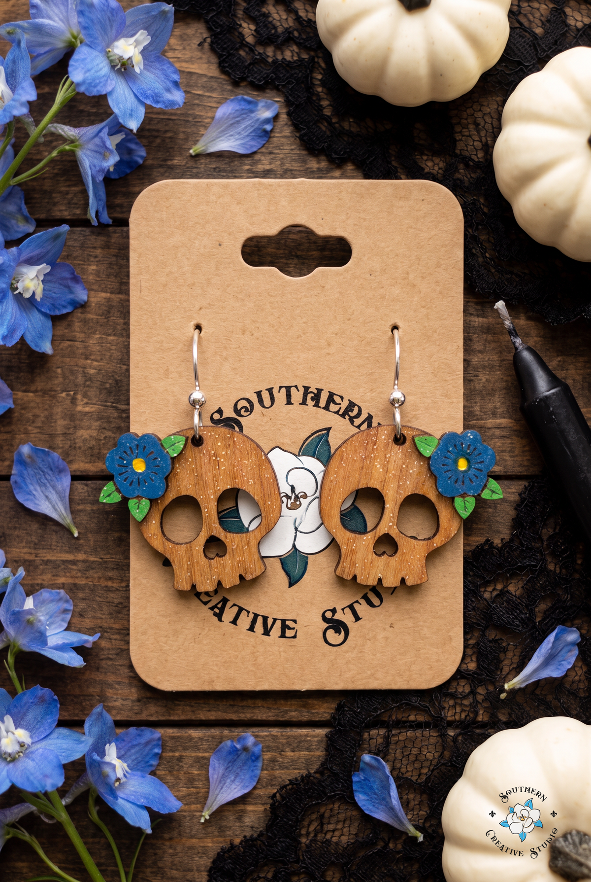 Flat lay of blue flower skull earrings on SCS card with delphinium, white pumpkins and lace