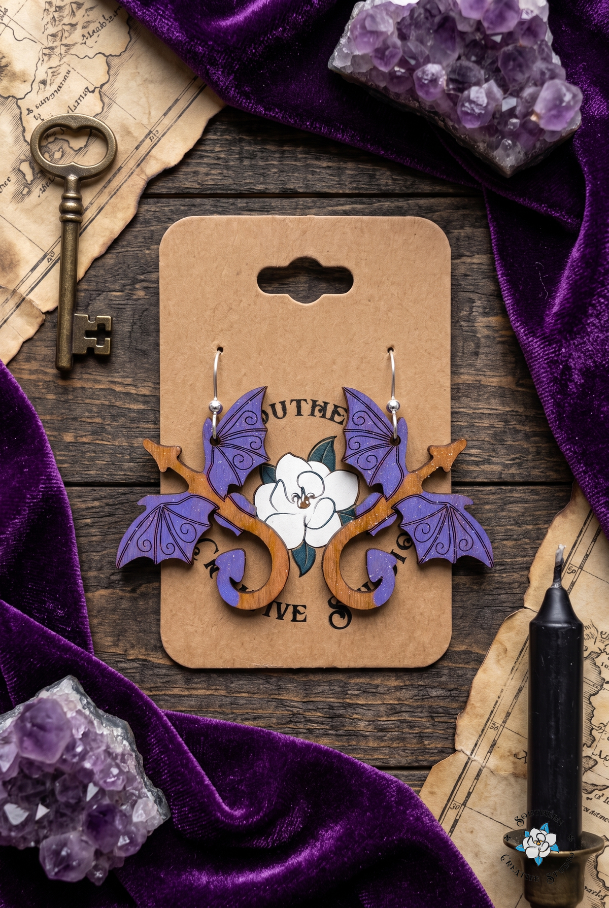 Flat lay of purple dragon earrings on SCS card with purple velvet, amethyst, brass key and parchment map