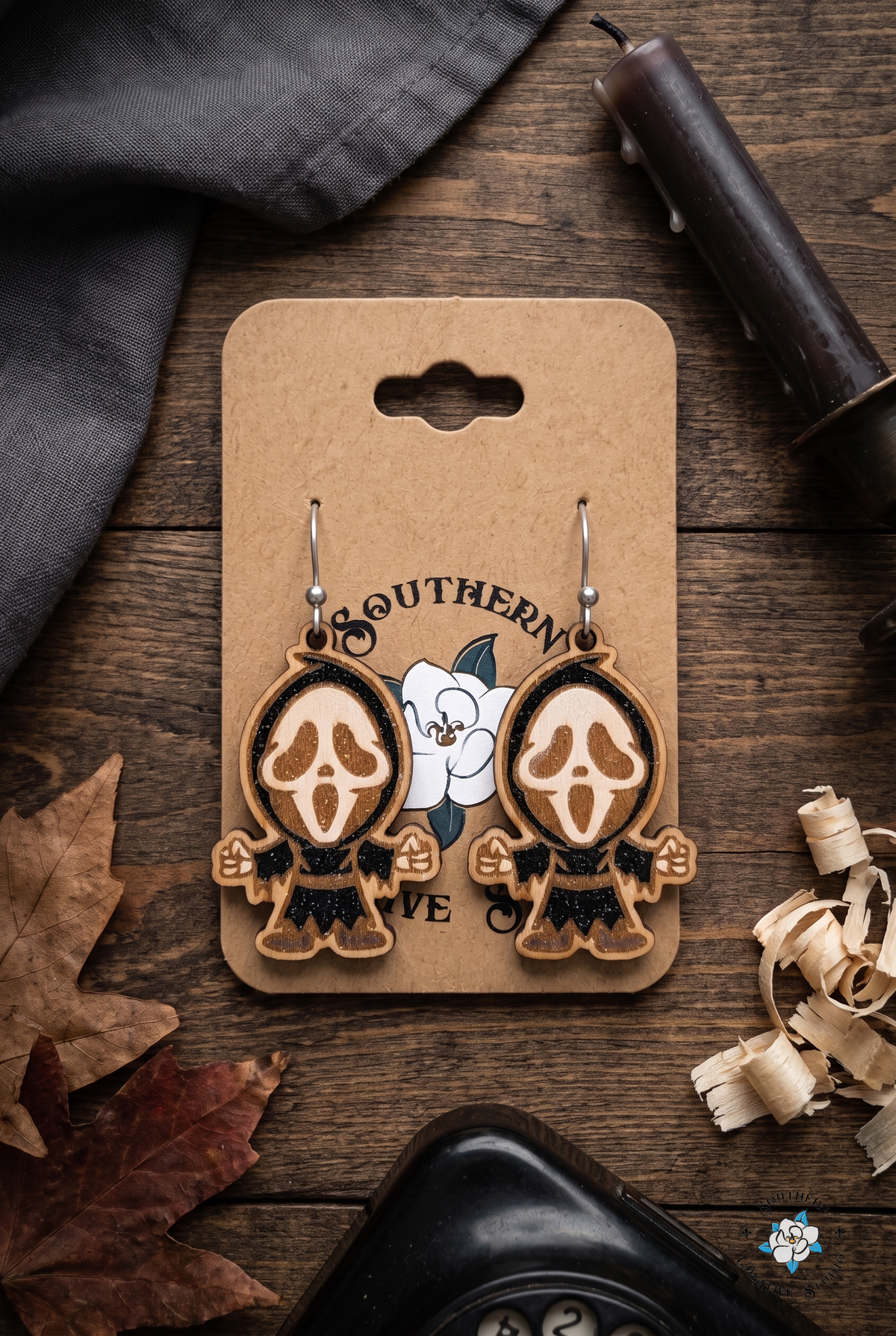 Moody flat lay with screaming ghost mask earrings on hang card, candle and autumn leaves