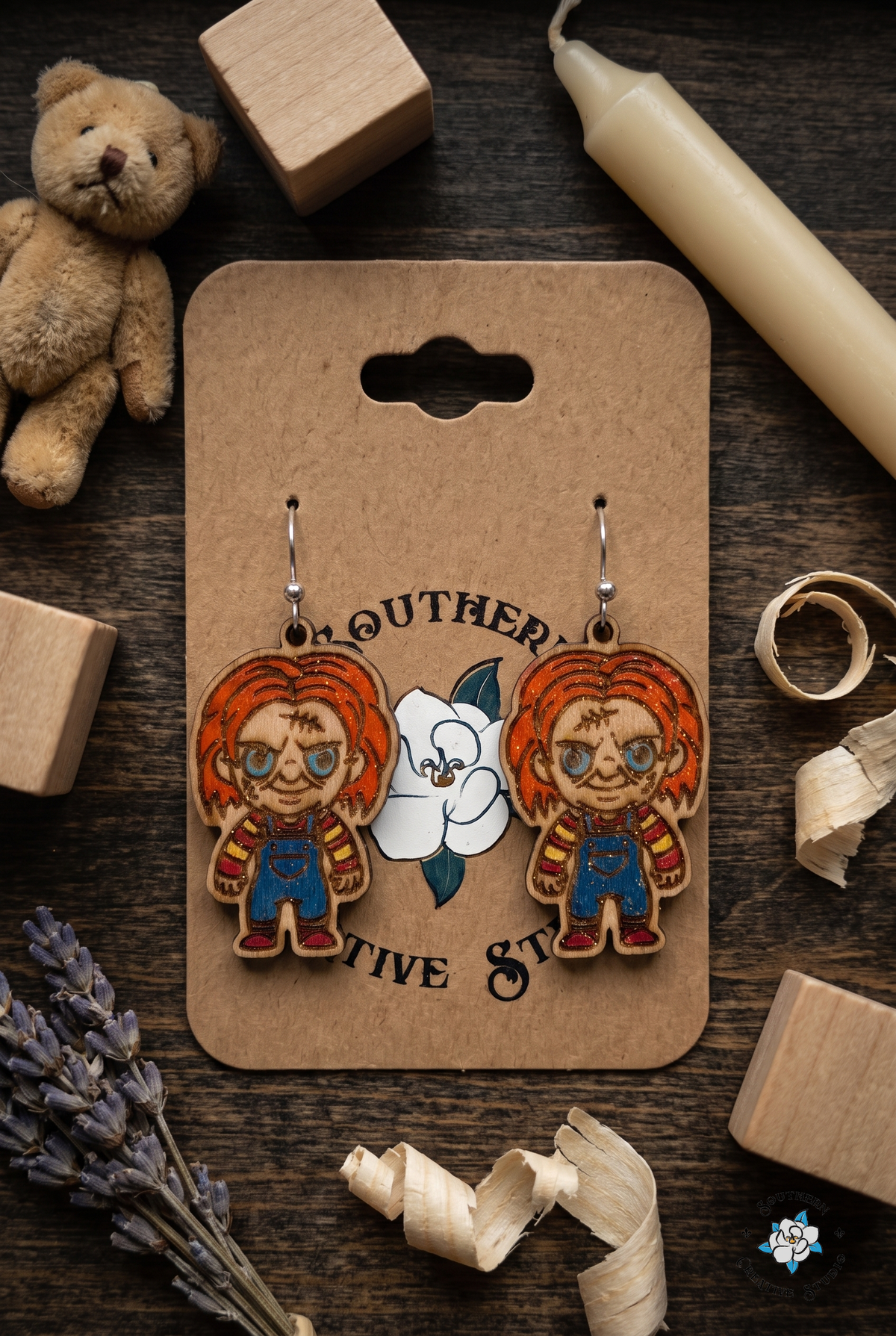 Creepy toy room flat lay with killer doll earrings on hang card, teddy bear and candle