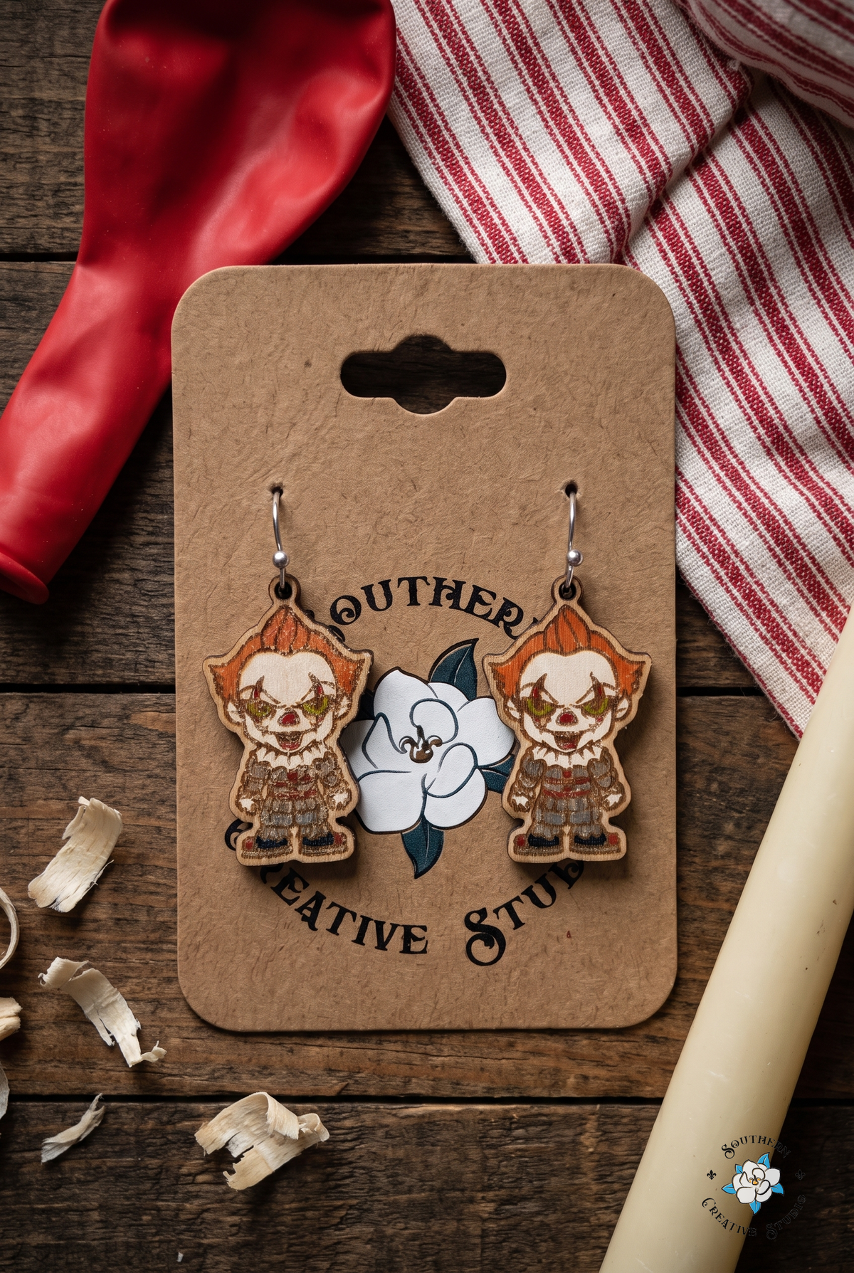 Carnival flat lay with creepy clown earrings on hang card, red balloon and candle