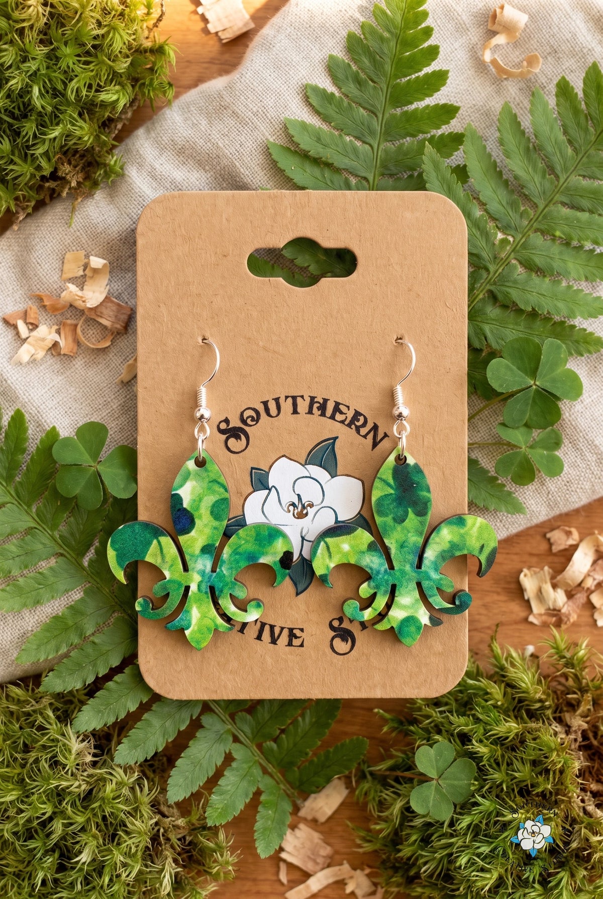 Fleur-de-lis clover earrings on card in a fern and shamrock flat lay