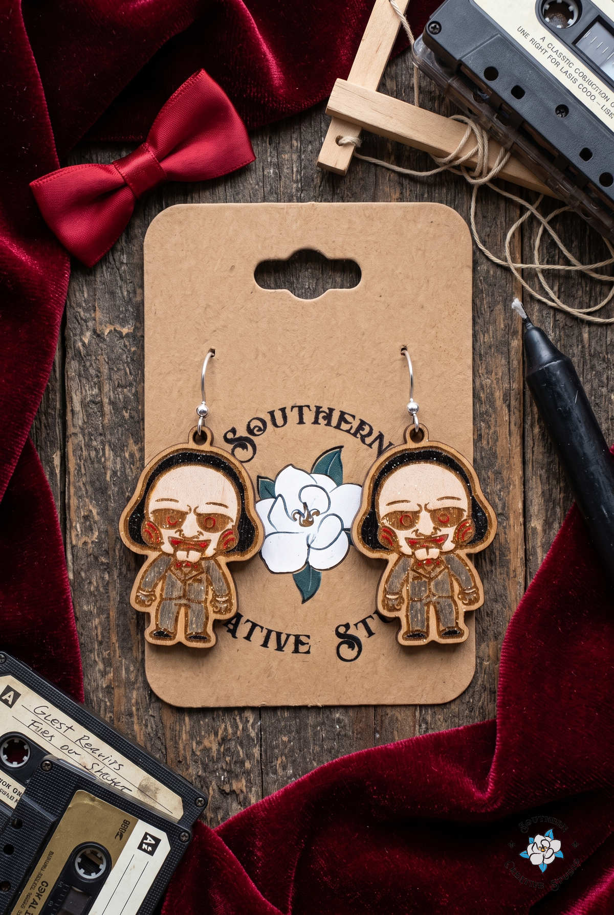 Flat lay of creepy puppet earrings on SCS card with red velvet, bow tie, cassette tapes and marionette bar