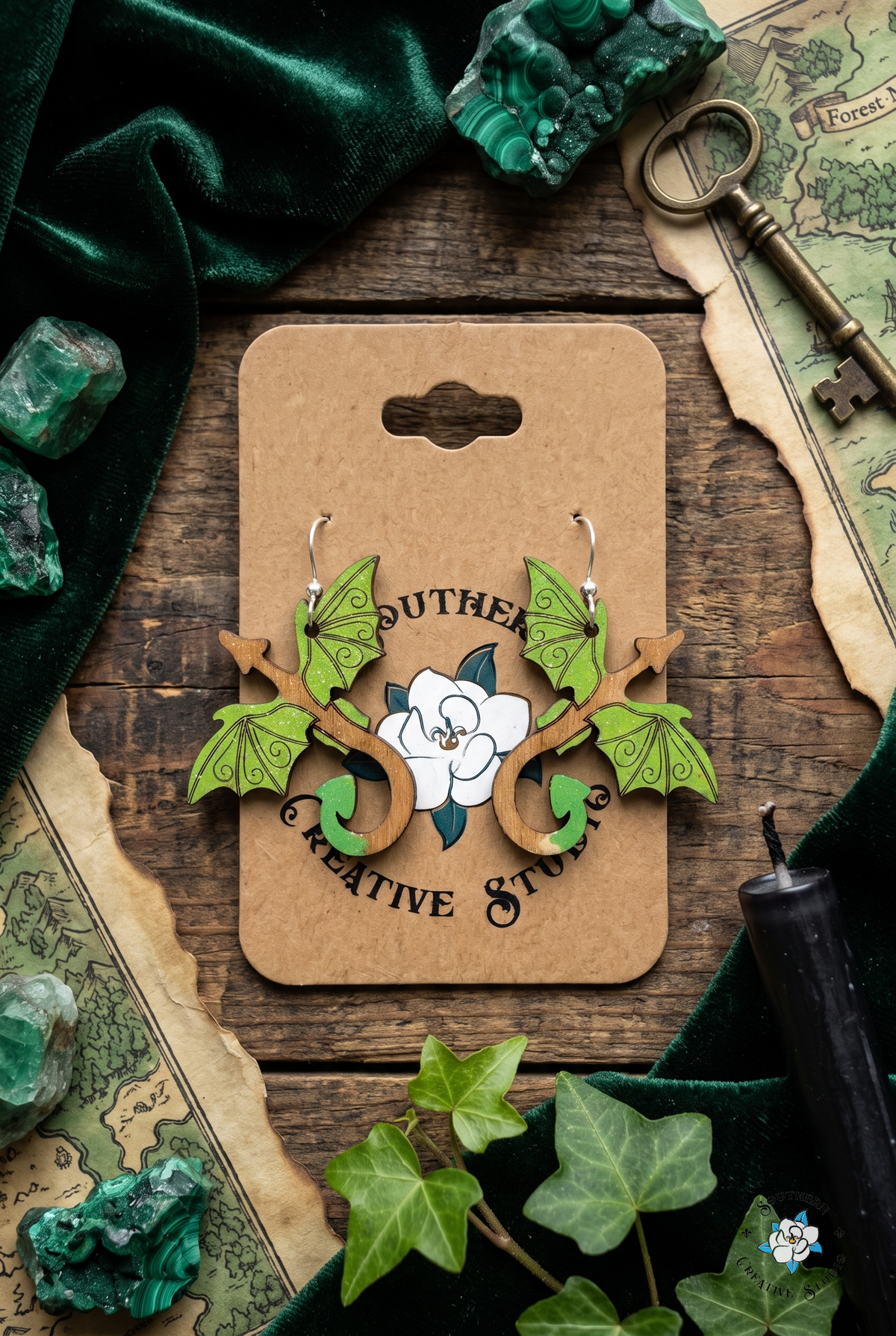 Flat lay of green dragon earrings on SCS card with green velvet, malachite, brass key, forest map and ivy