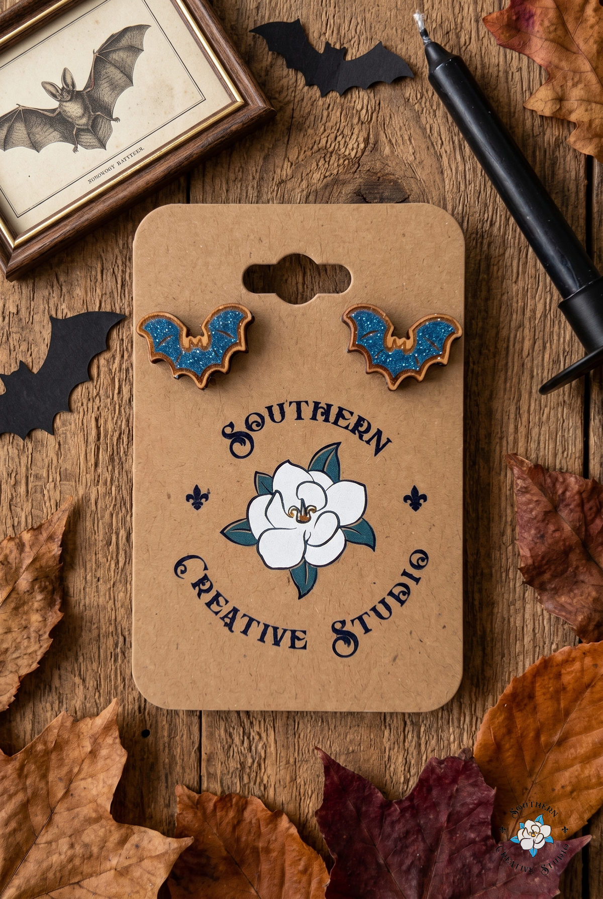 Flat lay of blue bat stud earrings on SCS card with bat print, paper bats and candle