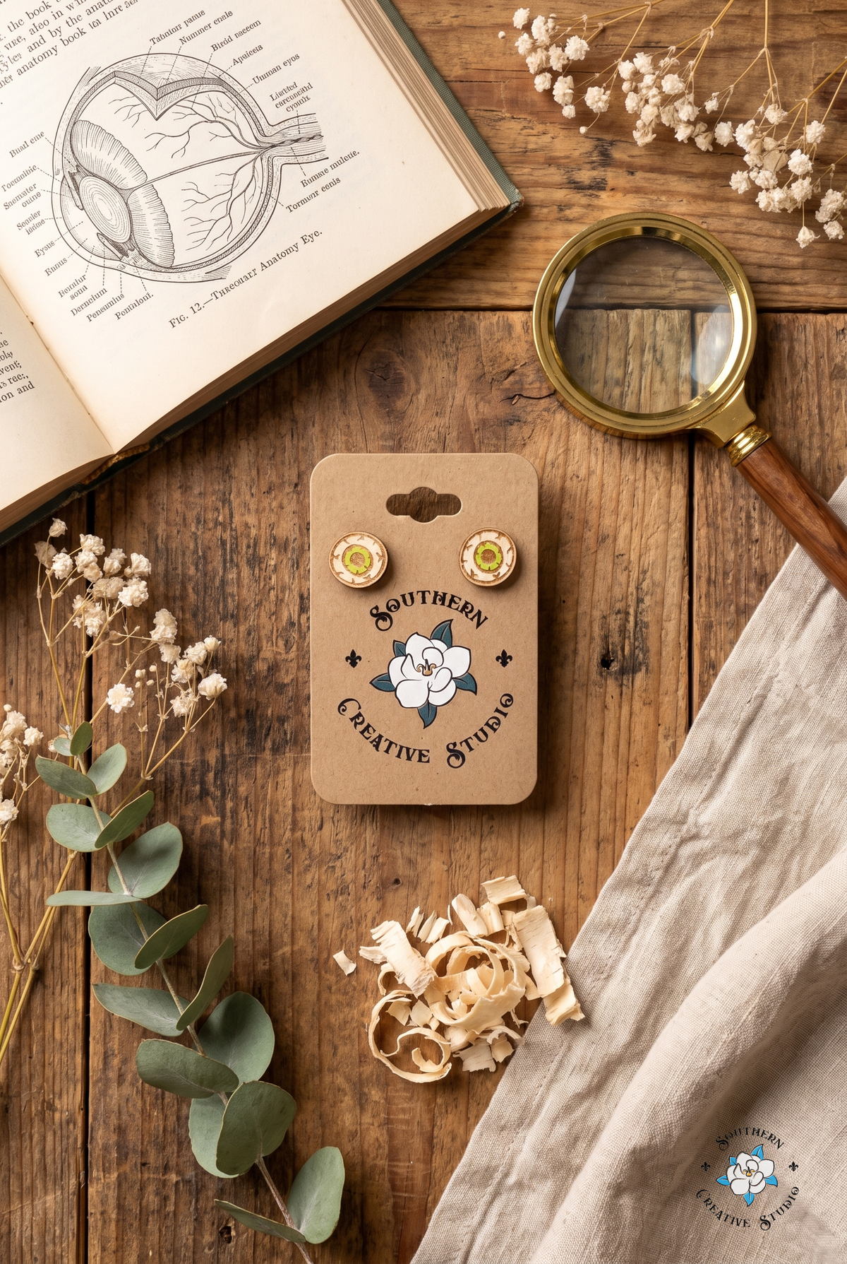 Green eyeball stud earrings on card flat lay with anatomy book and eucalyptus