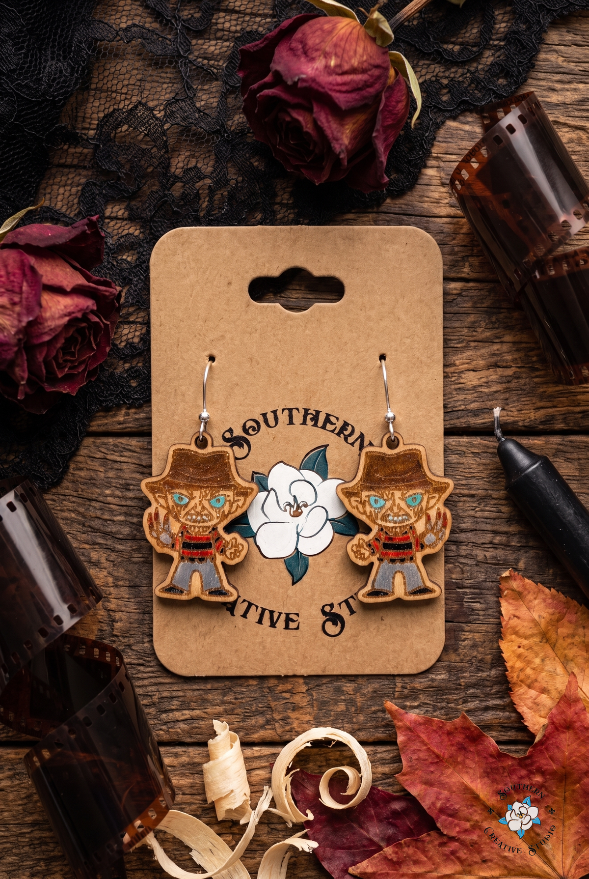 Spooky season flat lay of slasher wood earrings on SCS card with dried roses, lace, film strip and candle
