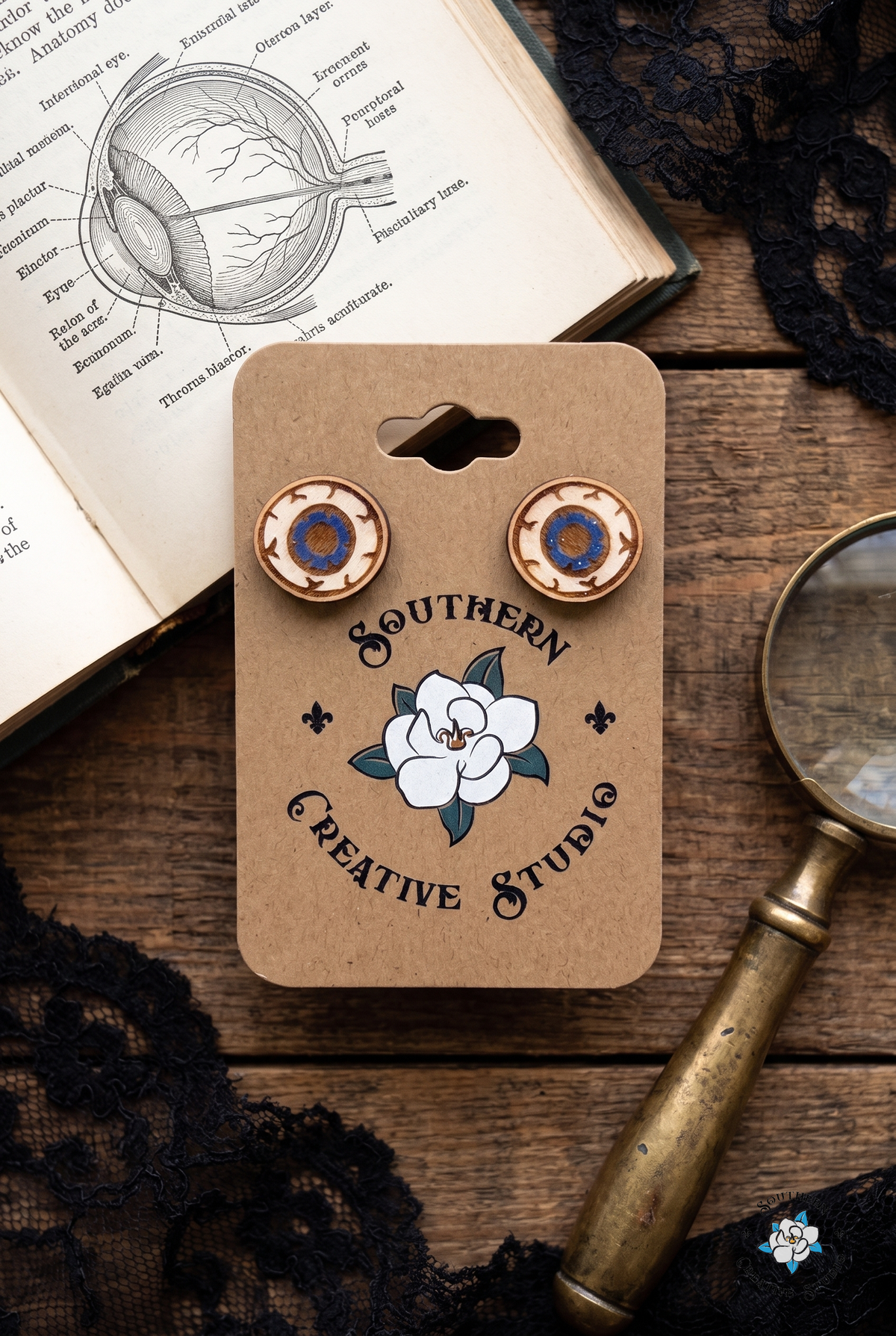 Flat lay of eyeball stud earrings on SCS card with anatomy book and magnifying glass