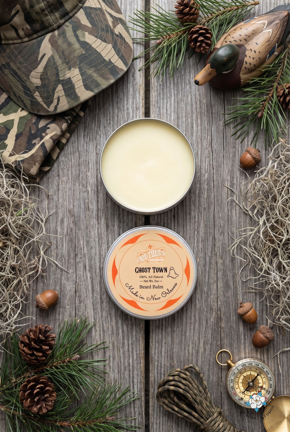 Ghost Town beard balm flat lay, open tin and lid with a misty Louisiana marsh with camo, a duck decoy and Spanish moss