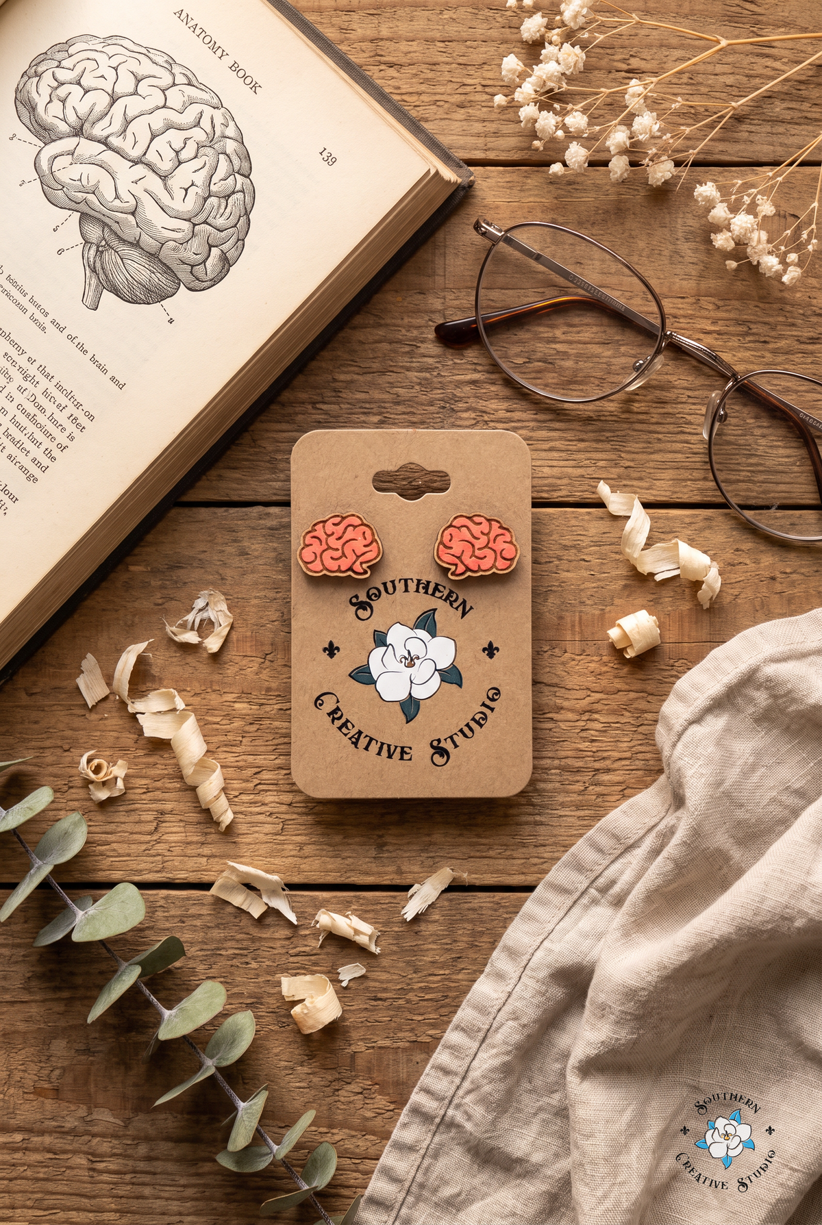 Pink brain stud earrings on card flat lay with anatomy book and eucalyptus